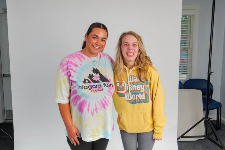 Two young women smile side by side, one in a tie-dye shirt and one in a yellow sweatshirt
