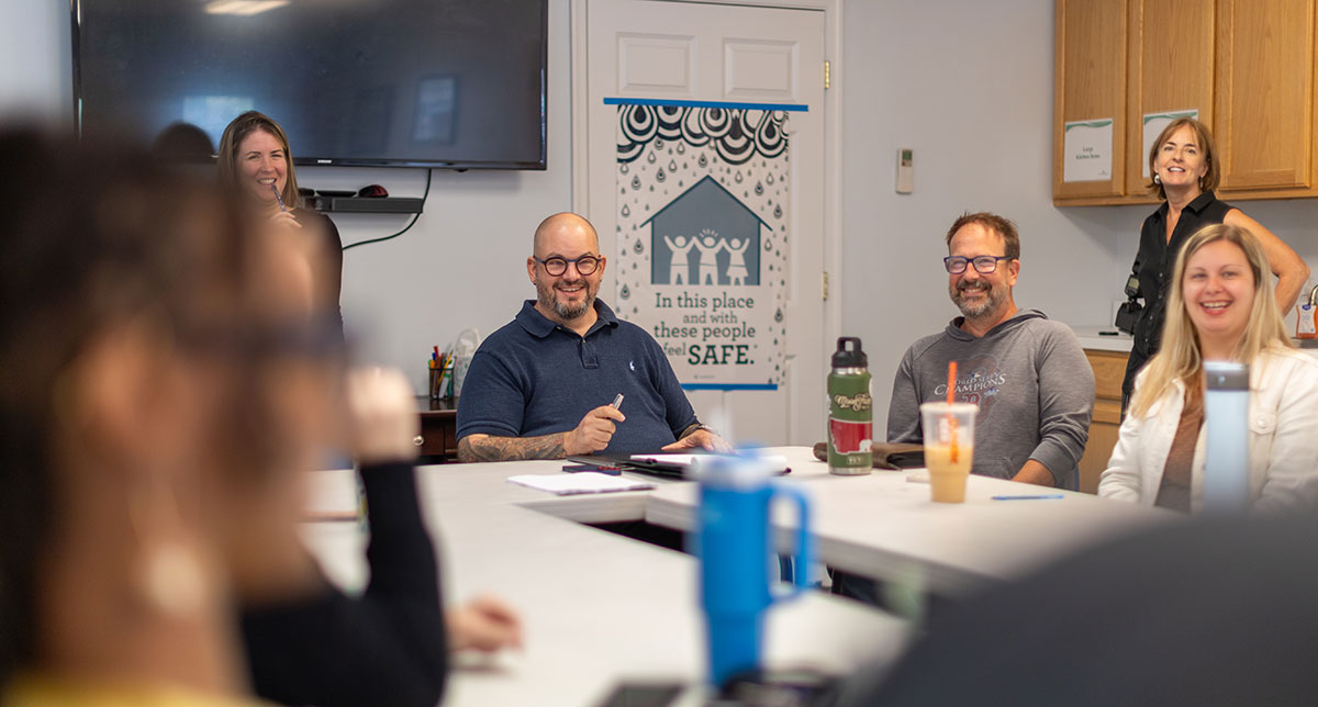 dr-matthew-ramsey-talking-to-community-living-managers-in-meeting A group of adults seated around a conference table during a meeting, smiling and engaged in conversation. A poster on the wall reads “In this place and with these people, I feel SAFE,” and additional staff members stand in the background near cabinets and a television.