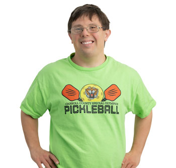 Target Community participant Jesse, wearing a green shirt that reads Carroll County Special Olympics Pickleball, brown hair and glasses, stands with his arms on his hips and smiling for camera.